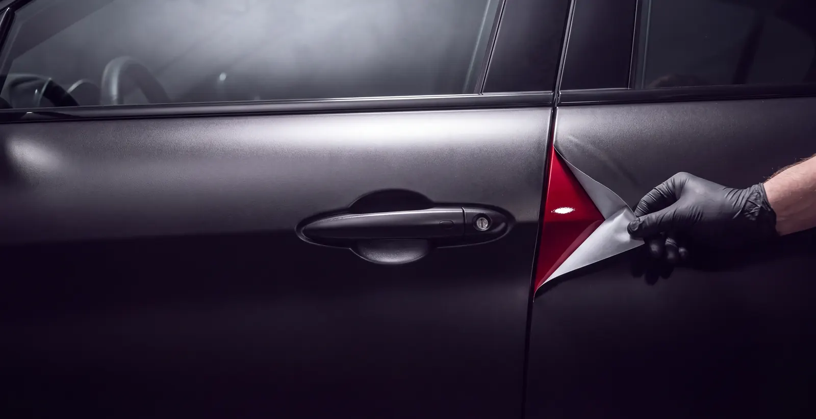 A gloved hand lifting the corner of satin black wrap on a car door, showing glossy red paint underneath
